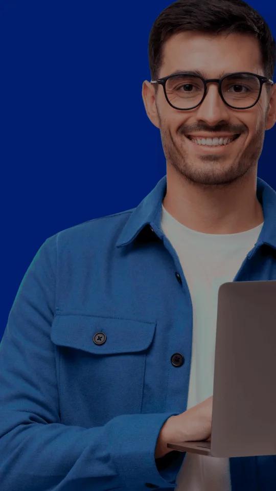 Homem branco de cabelo escuro e barba rala usando óculos de armação preta sorri para a câmera. Ele está de pé com uma camisa azul clara aberta e usa camiseta branca. Ele segura um notebook com as mãos.