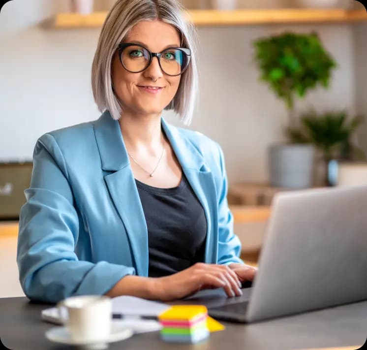 Empreendedora sorri para a câmera enquanto mexe no notebook. Ela possui cabelos curtos e está usando óculos de grau, camisa preta e blazer azul claro.