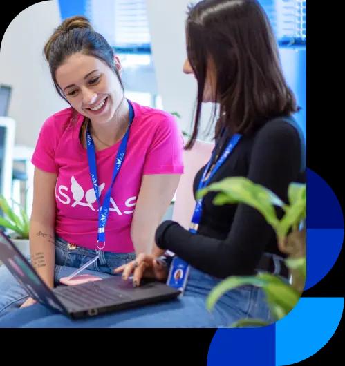 Colaboradoras do Asaas trabalham na sede. Uma delas, à esquerda, sorri enquanto olha para o notebook. A outra, à direita, está de perfil, com o notebook no colo.