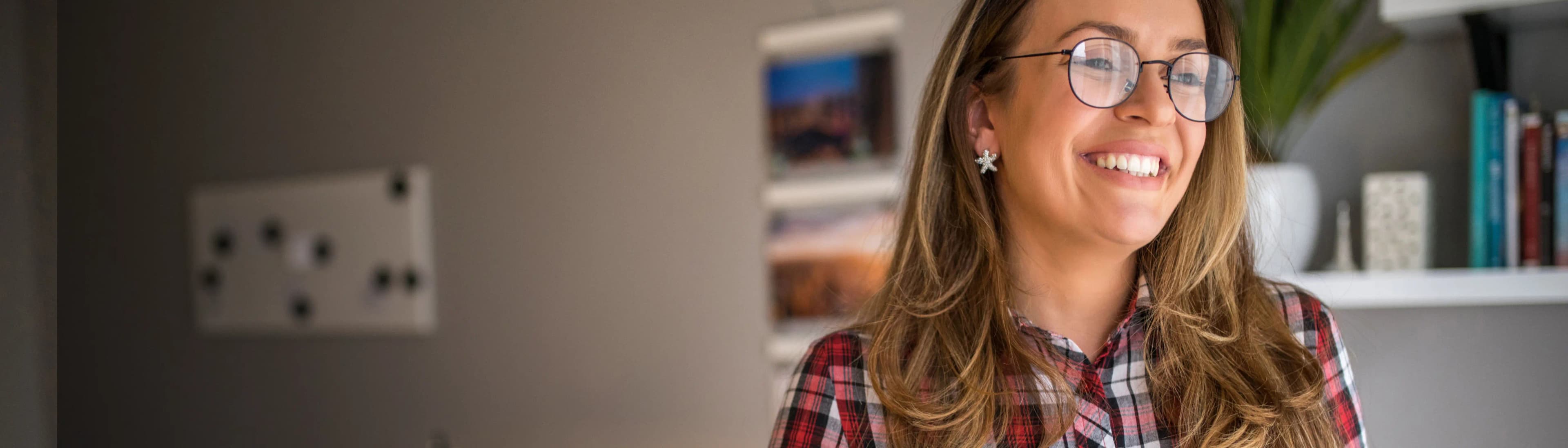 Empreendedora sorri, enquanto olha para sua direita. Ela tem pele clara e cabelos lisos, com tons castanhos e acobreados. Também veste uma camisa de botão xadrez, nas cores verde e marrom. Ela segura um tablet com a mão direita, junto ao corpo.