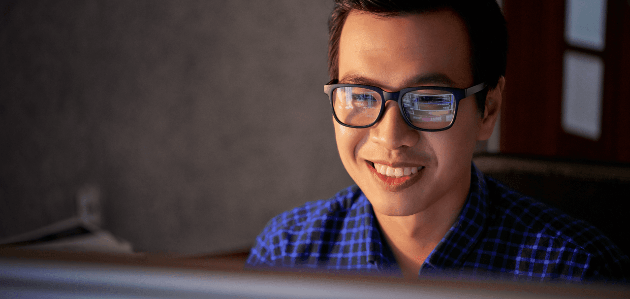 Desenvolvedor sorri para a tela do computador enquanto usa a API de pagamentos do Asaas. Ele tem a pele clara, cabelos curtos e castanhos, e usa óculos de grau com armação preta. Além disso, veste uma camiseta xadrez na cor azul. Ao fundo, há uma parede cinza e uma janela.