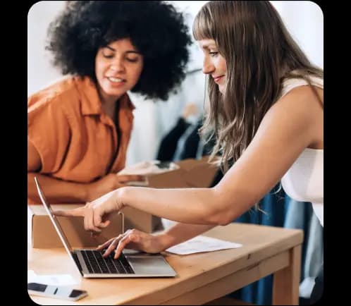 Duas mulheres trabalhando juntas em uma mesa. Uma delas aponta para a tela de um notebook enquanto a outra observa.